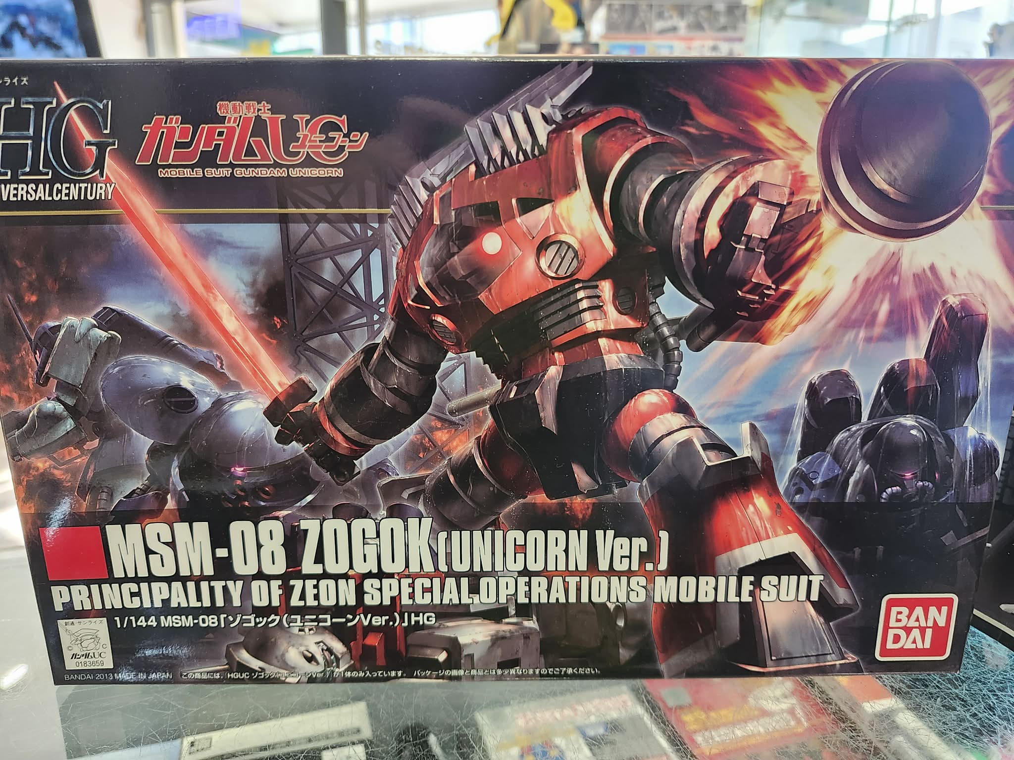 Photographie de la boîte de maquette Bandai Namco High Grade représentant le mobile suit MSM-08 Zogok dans sa version issue de Mobile Suit Gundam Unicorn, en action avec ses armes.