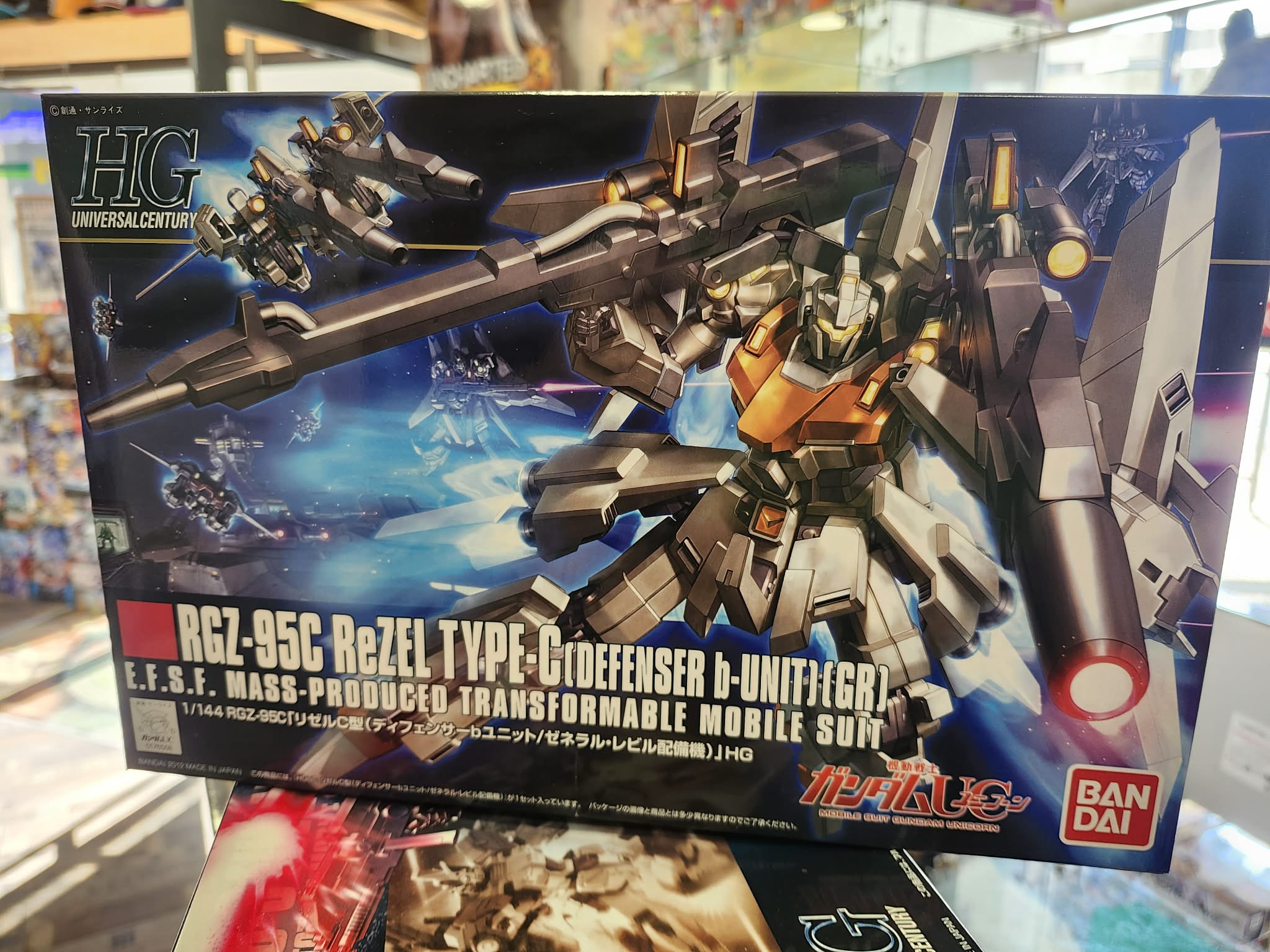 Photographie de la boîte de maquette Bandai Namco High Grade (HG) représentant le mobile suit transformable RGZ-95C ReZEL Type-C (Defenser b-Unit) (GR) issu de l'Universal Century, présenté en action avec ses équipements lourds.